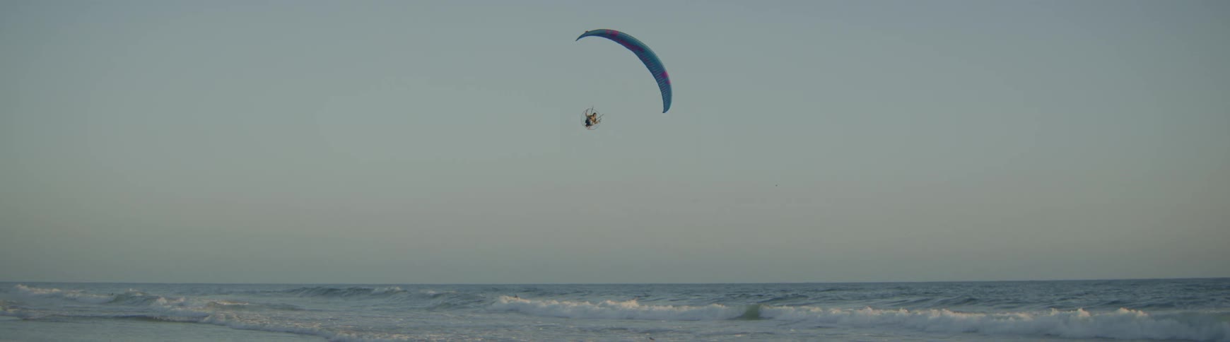 A lone paramotor pilot glides serenely above the ocean waves under a vast, clear sky