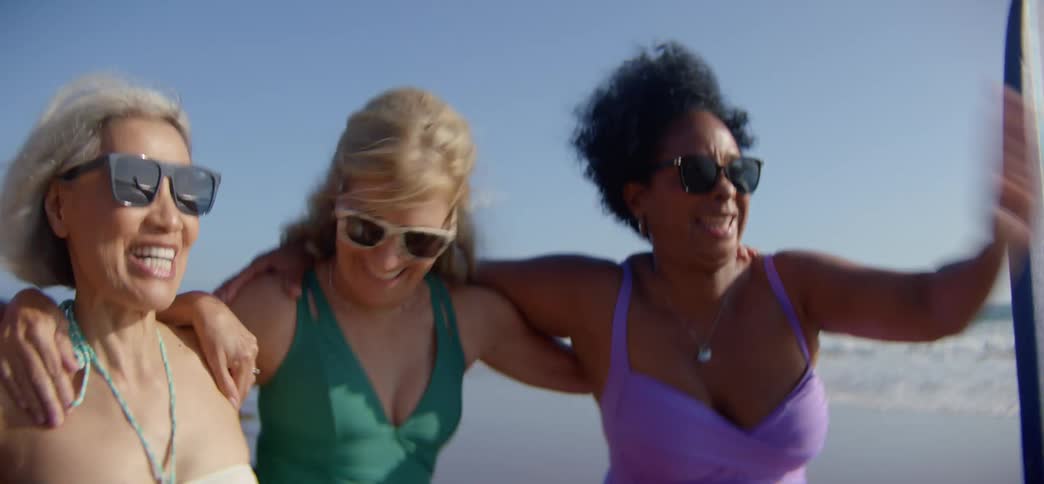 A diverse group of cheerful senior women in swimsuits walk arm-in-arm on a sunny beach, laughing and celebrating their friendship in slow motion