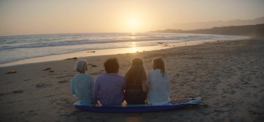 A diverse group of four women friends sit together on a surfboard on the beach, watching the beautiful sunset over the ocean