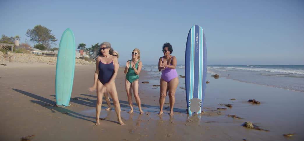 A group of diverse and joyful senior women in swimsuits dance energetically on the beach with their surfboards in slow motion
