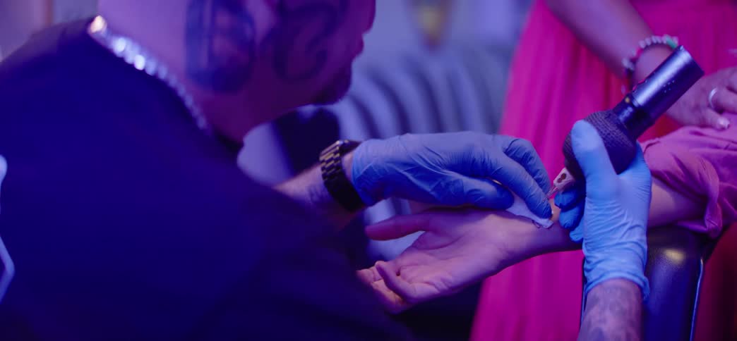 A happy senior woman laughs and grimaces as a tattoo artist gives her an infinity symbol tattoo on her wrist at a party