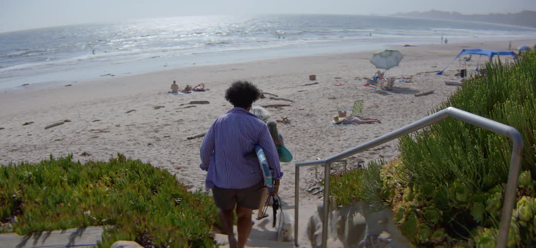 A diverse group of older women friends with surfboards and a dog walk down a sandy path to the ocean for a day at the beach