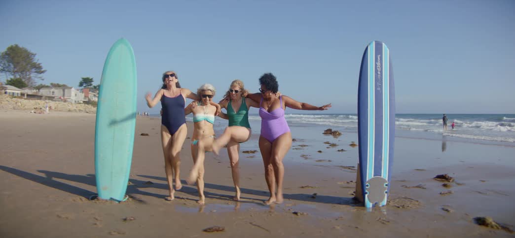 Four joyful senior women in swimwear have fun performing a synchronized dance on the beach between their surfboards