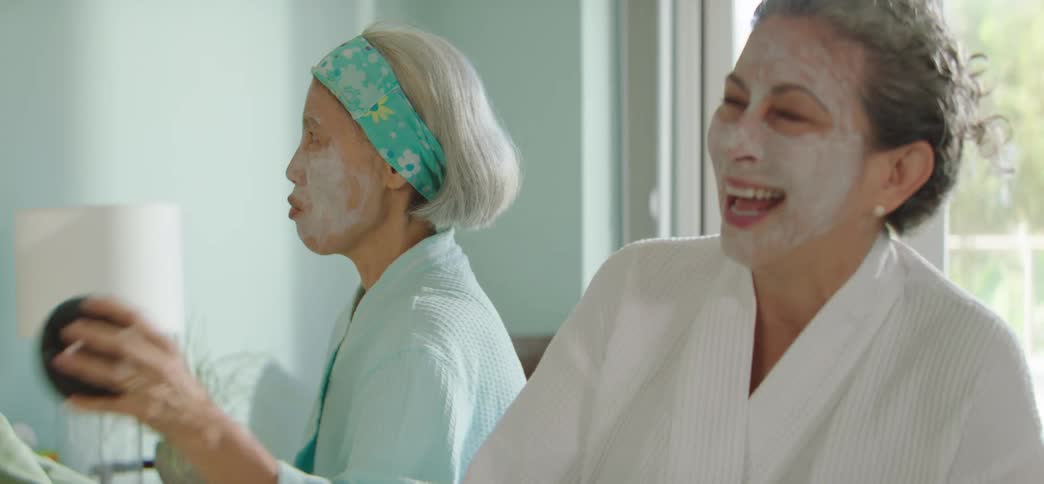 Two cheerful elderly female friends with facial masks dance and laugh while listening to music during a spa day at home