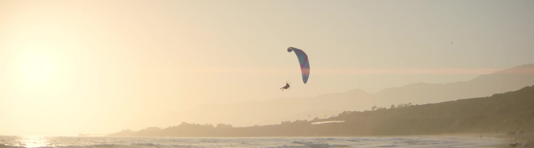 A person on a powered paraglider soars peacefully over the ocean coastline during a hazy sunset