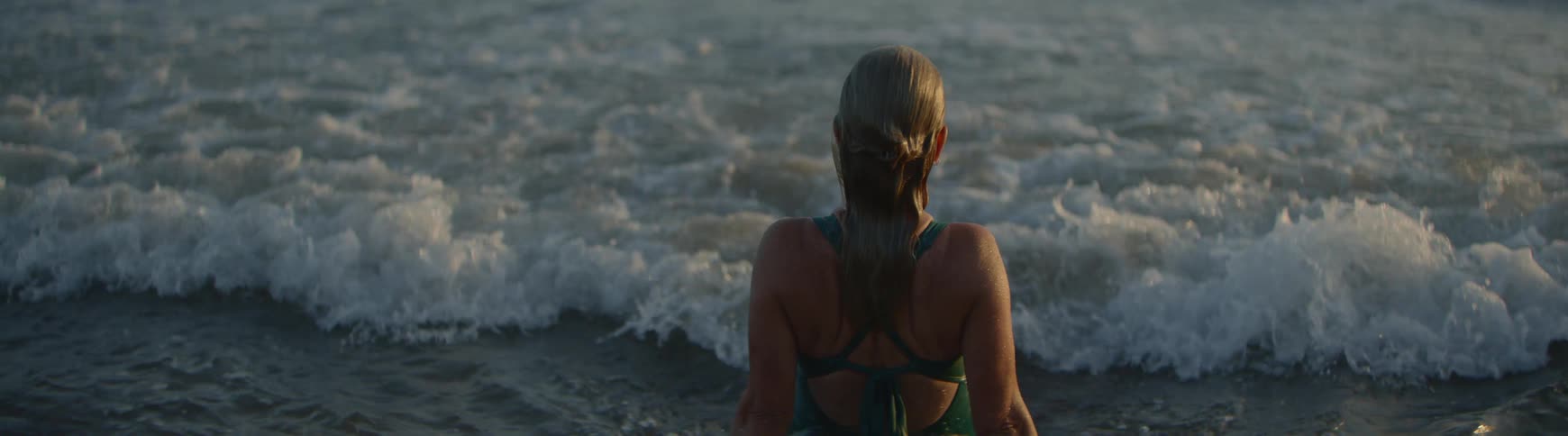 A woman with her back to the camera sits in the shallow ocean water, looking out at the gentle waves in slow motion