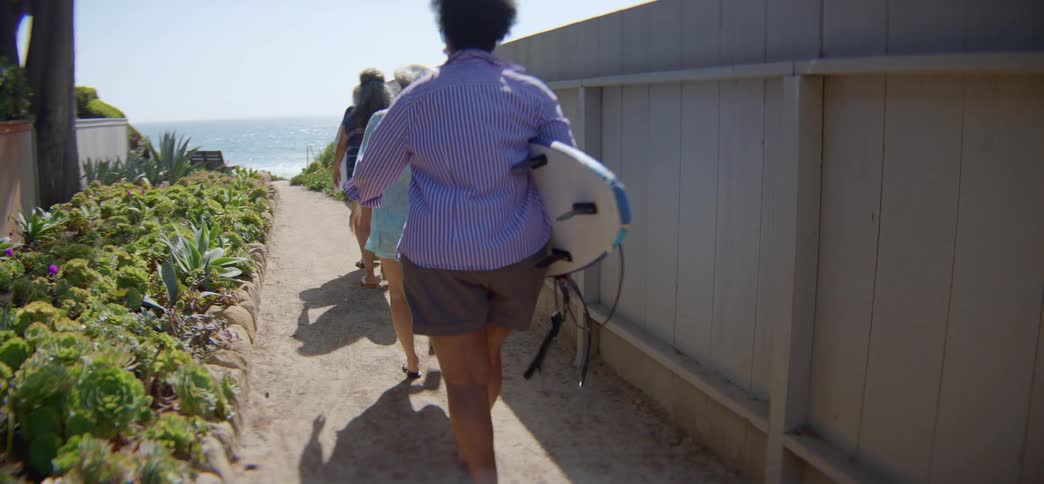 A group of senior women friends walk down a sunny, sandy path with their surfboards, heading to the ocean for a day of surfing