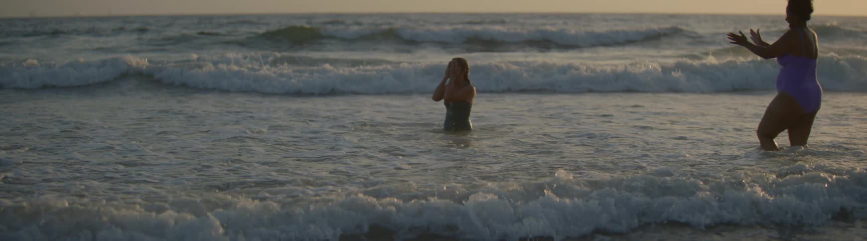 Two happy female friends playfully splash each other while swimming in the ocean at sunset in slow motion