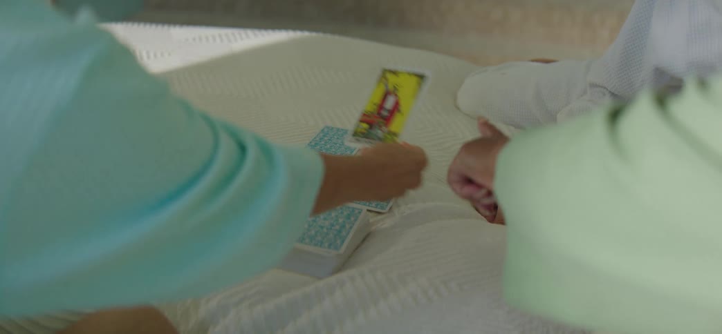 A woman in a teal robe performs a tarot card reading for a friend on a white bedspread in slow motion