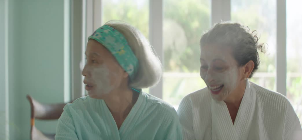 Elderly women in bathrobes and face masks enjoy a spa day at home, laughing as one reads tarot cards on the bed