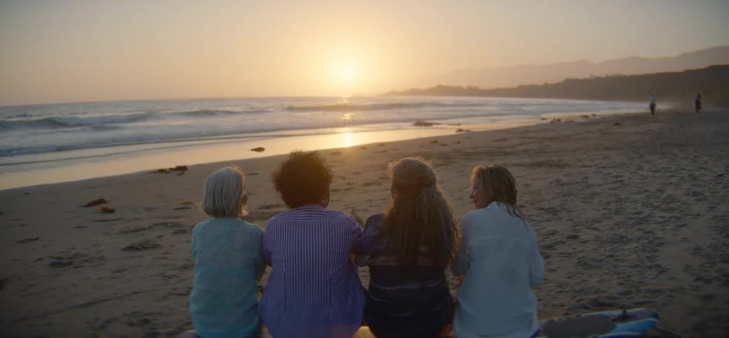Four diverse senior women sit together on a surfboard on the beach, watching the golden sunset over the ocean