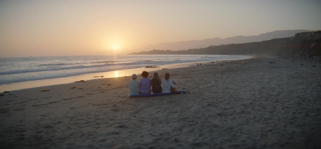 Four friends sit together on a surfboard on the sand, peacefully watching the sun set over the tranquil ocean waves