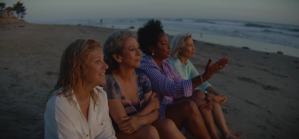 A diverse group of four senior women friends sit together on the sand, talking and pointing at the ocean as they enjoy a beautiful sunset