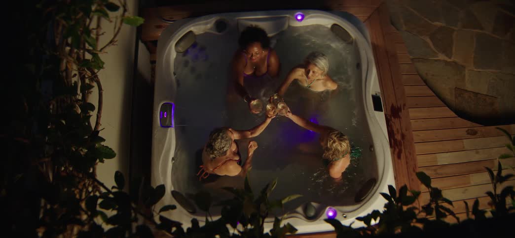 A group of diverse, mature women share drinks and make a toast while relaxing together in a hot tub at night