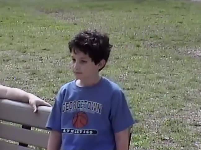 A young boy in a blue shirt standing by a park bench suddenly gasps in shock and points at something off-camera