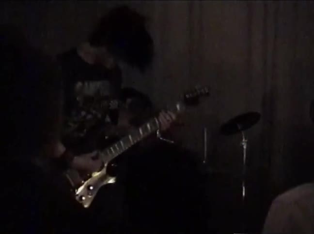 Grainy, low-light footage captures a rock band guitarist with long hair headbanging energetically during a live performance