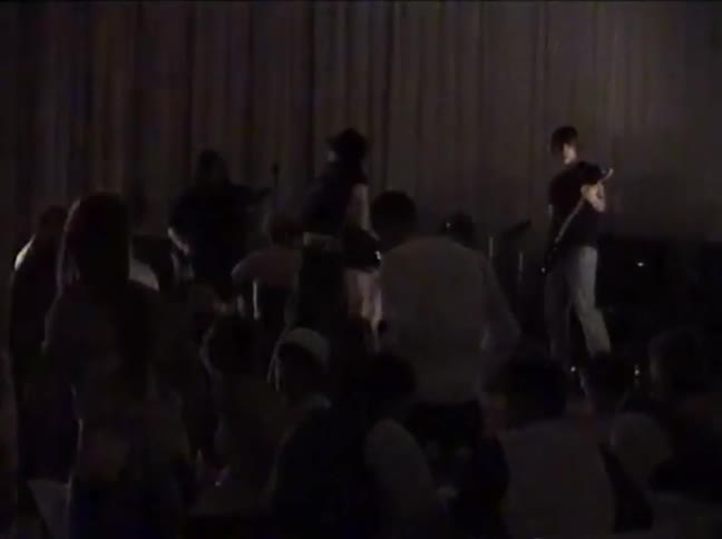 A young rock band performs for a crowd on a dimly lit stage in grainy, vintage-looking footage
