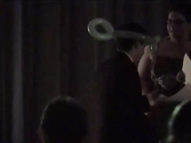 A man in a suit with a silly key-shaped balloon on his head at a dimly lit party, captured in grainy, vintage-style footage