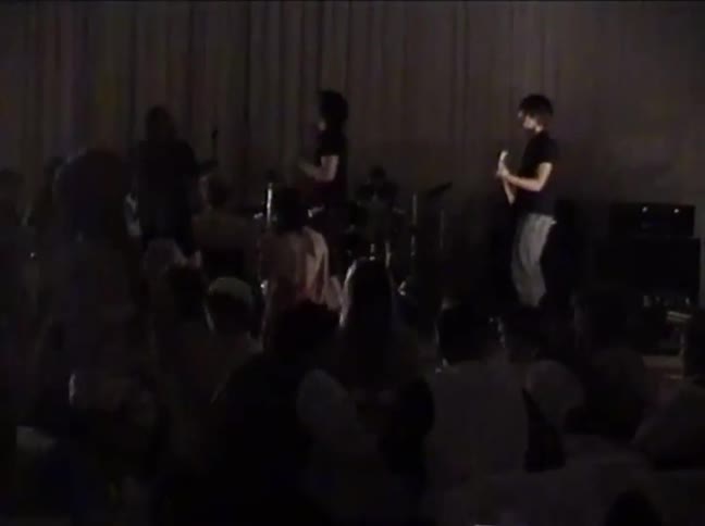 Vintage, grainy footage shows a band performing on a dimly lit stage for an enthusiastic audience