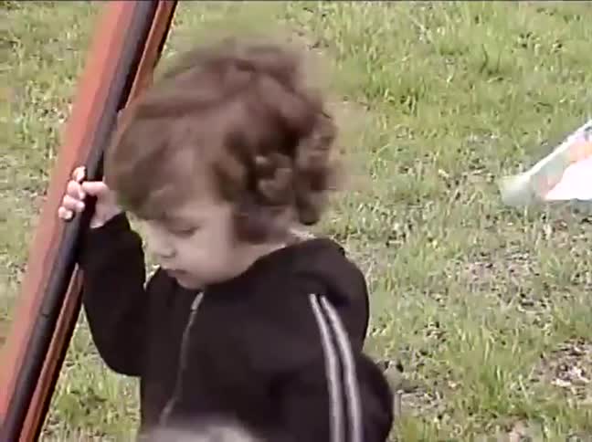 A cute curly-haired toddler holds onto a pole during a family outing at a park in this vintage home video