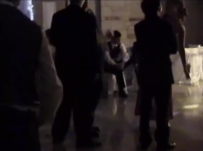 Grainy, low-light vintage footage shows silhouettes of teenagers in formal wear standing around at a school dance