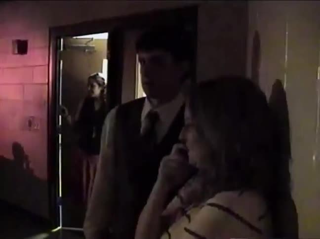 A young man and woman dressed for a formal event chat in a dimly lit corridor in this retro home video from the 1990s