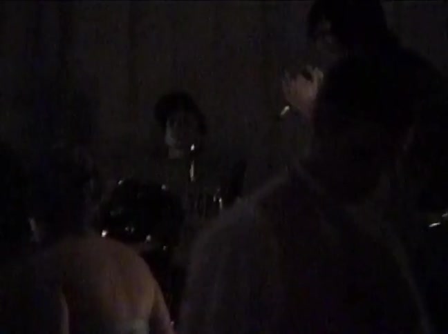 Grainy, low-light footage shows a band performing an energetic set for a crowd in a dark club