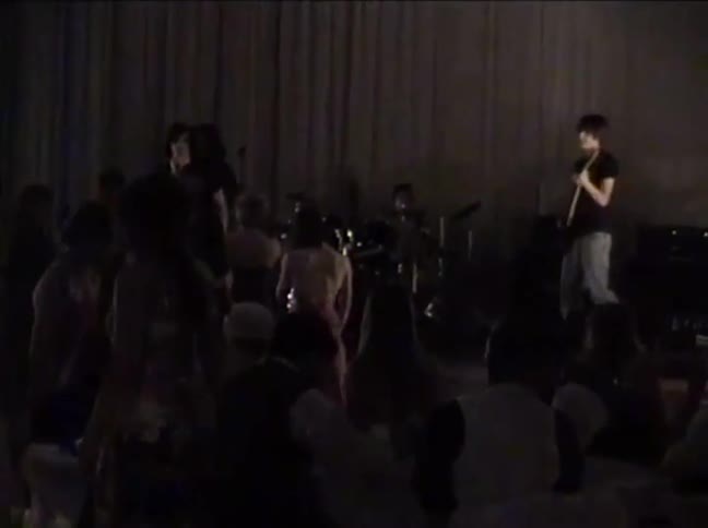 Vintage low-quality footage shows a rock band performing a live concert for an audience in a dark venue