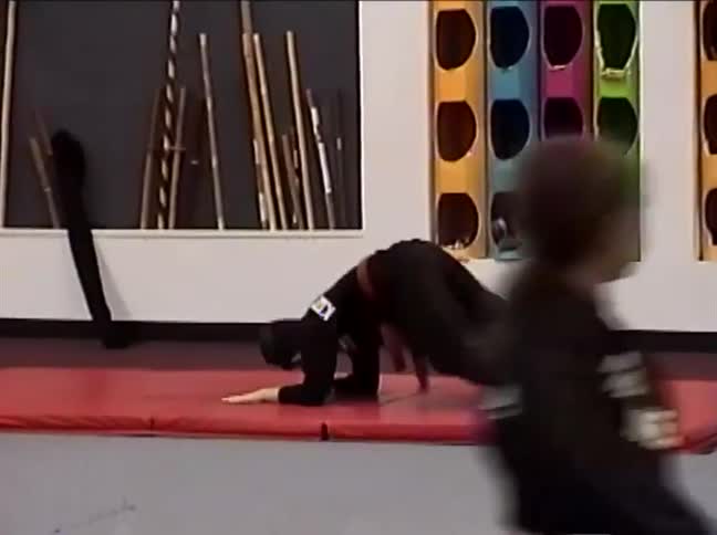 A young boy in a black gi and red belt performs a complex tumbling and martial arts routine on a red mat at a competition