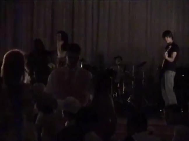 A rock band performs on a dimly lit stage for an audience in this vintage VHS-style footage