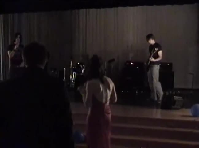 Lo-fi vintage footage shows a young rock band performing on a dimly lit stage as the audience watches