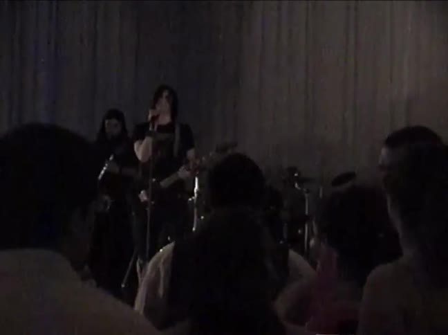 A punk rock band performs on a dimly lit stage for an audience in this grainy, vintage-style footage