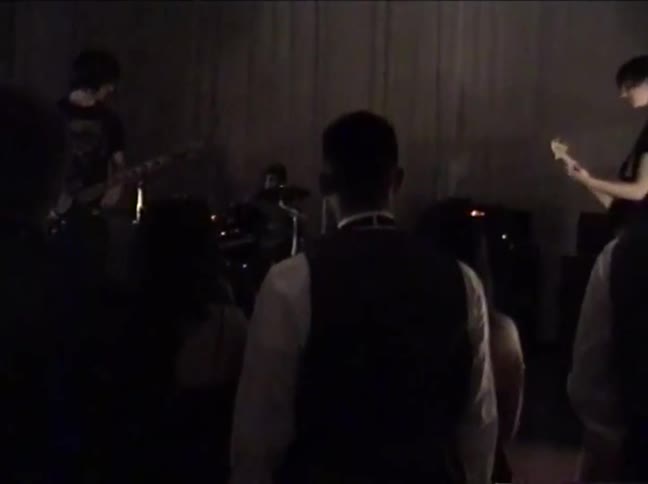 Vintage, low-light home video footage shows a rock band performing on stage from behind the audience