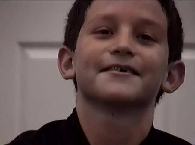 A happy young boy with a gap-toothed smile and braces looks up in a vintage home video