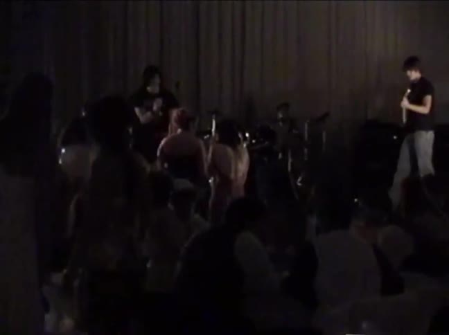 Grainy vintage footage shows a rock band performing live on a dark stage for a crowd