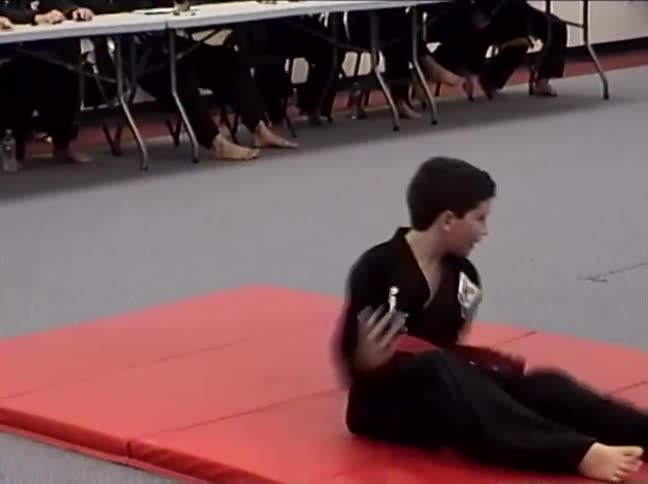 A young martial artist with a red belt demonstrates various falling and rolling techniques on a red mat in a dojo