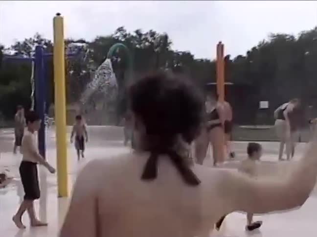A vintage home video shows children and adults playing in the sprinklers at a crowded splash pad on a summer day