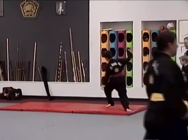 A young boy in a black uniform demonstrates an acrobatic martial arts form on a red mat during a test