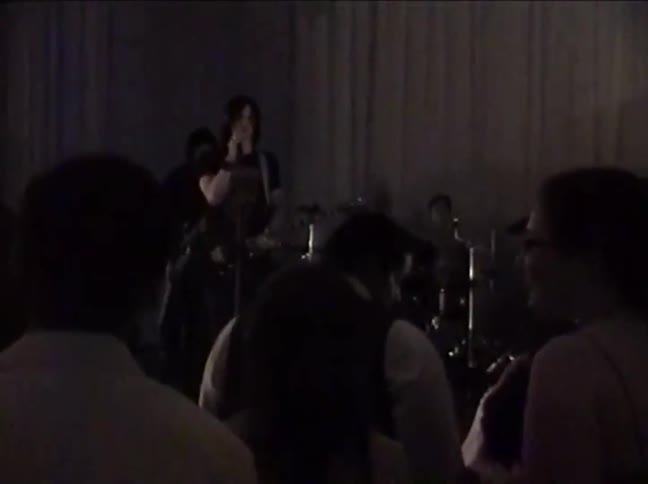 Grainy, vintage VHS footage of a rock band performing live on a dimly lit stage for an audience