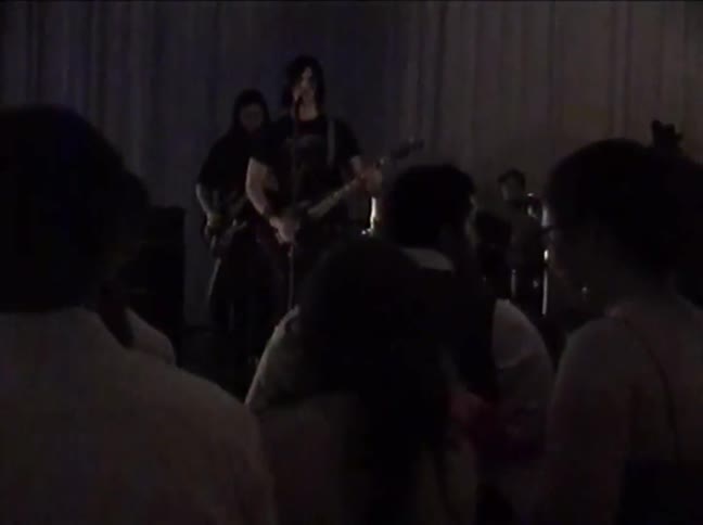 Vintage, low-quality footage shows a rock band performing a concert for an energetic crowd in a dimly lit venue