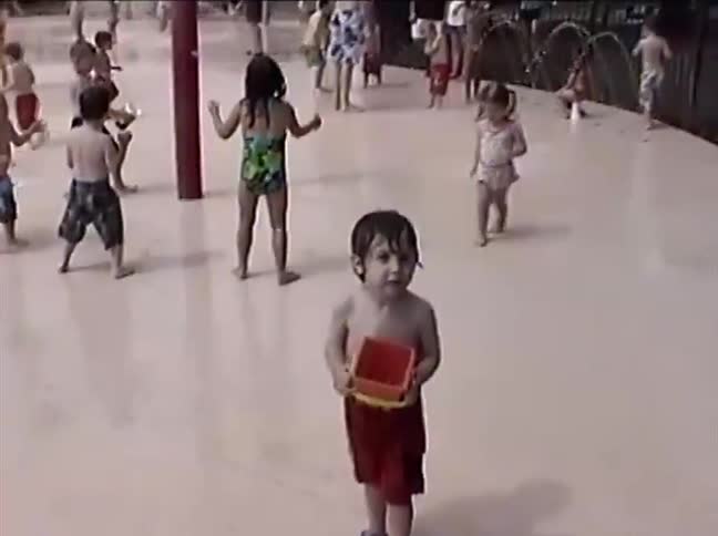 A young toddler boy in swim trunks holds a small bucket while playing at a crowded splash pad on a summer day