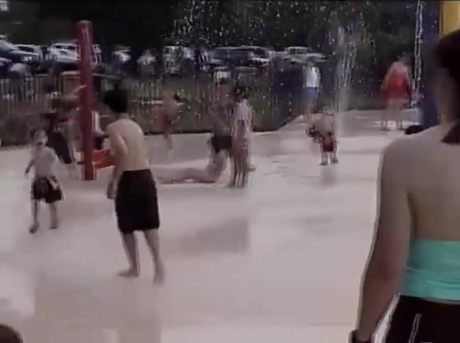 Low-quality vintage footage shows children and adults having fun and cooling off at a public splash park on a hot summer day