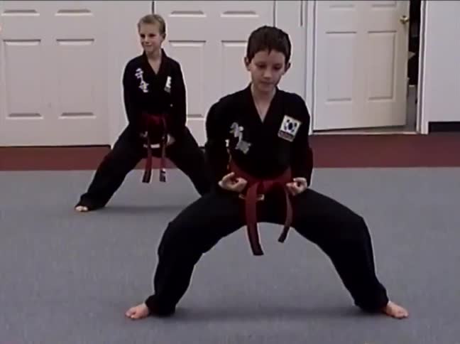 Two young boys in black uniforms and red belts practice a slow, synchronized martial arts kata in a dojo