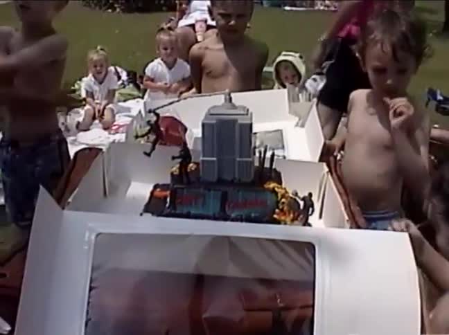 Children at a sunny outdoor birthday party excitedly gather around a large, superhero-themed cake