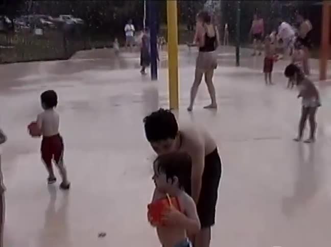 Grainy home video footage captures a nostalgic family day out as children and adults play in the sprinklers at a splash park
