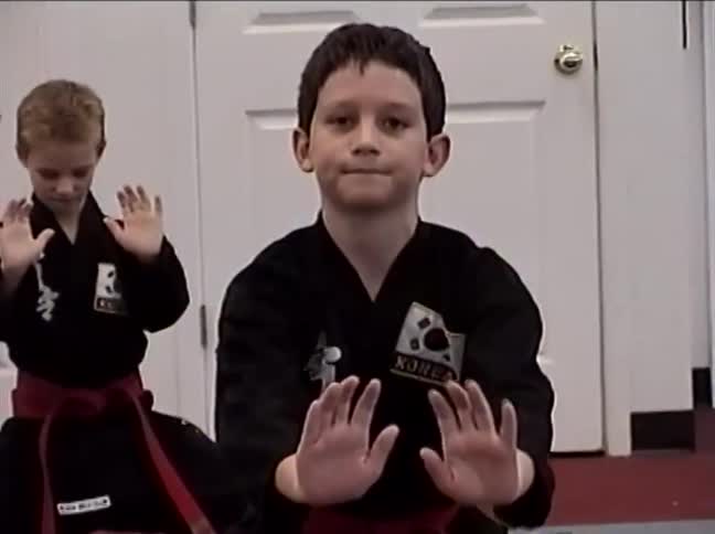 A young boy in a black gi and red belt tries to meditate seriously during his martial arts class before breaking into a toothy smile