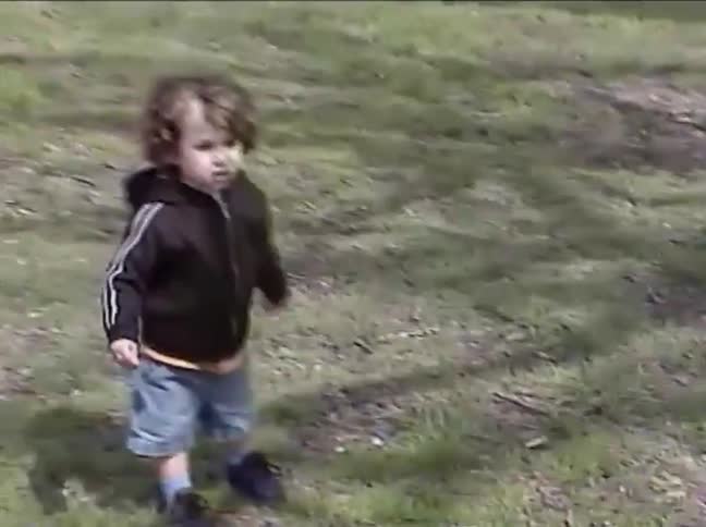 Vintage home video footage shows a cute toddler with curly hair walking across a grassy park with other children and an adult