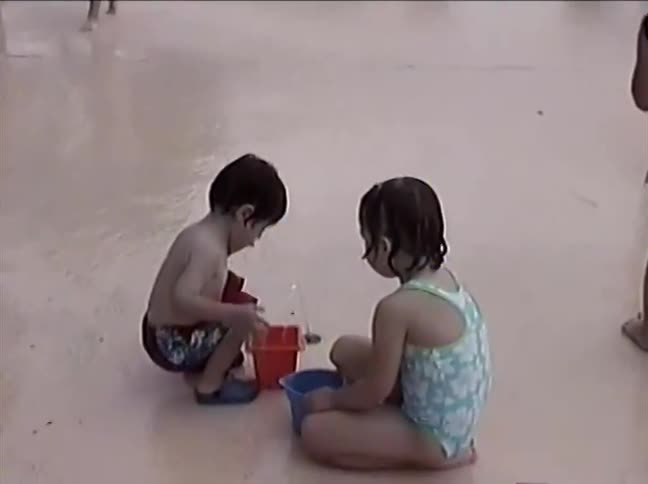 A group of children in swimsuits play joyfully at a splash pad in this grainy, vintage-style home movie