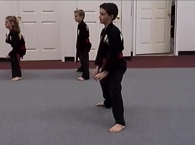 A group of young children in black uniforms practice synchronized martial arts kicks and stances in a dojo