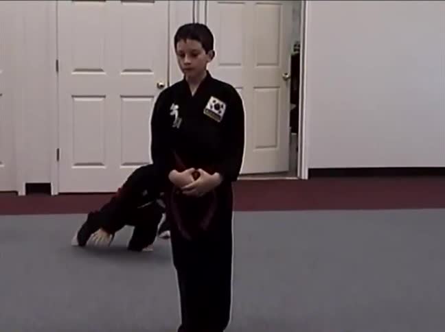 A young boy in a black dobok with a red belt practices a complex martial arts form in a dojo as another child practices in the background